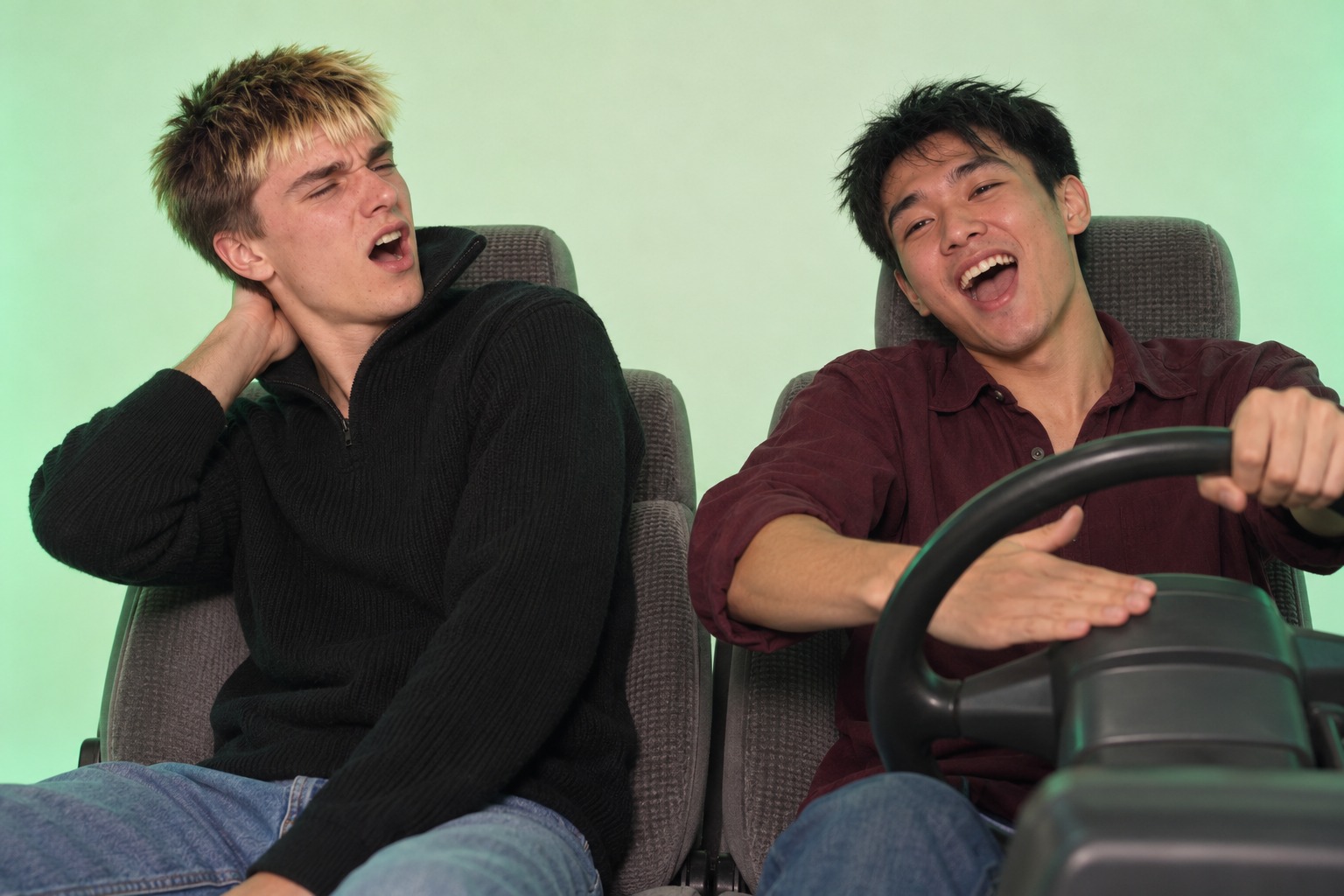 Two young men in boxy car seats sing badly, one laughing at the wheel and one flinching away.