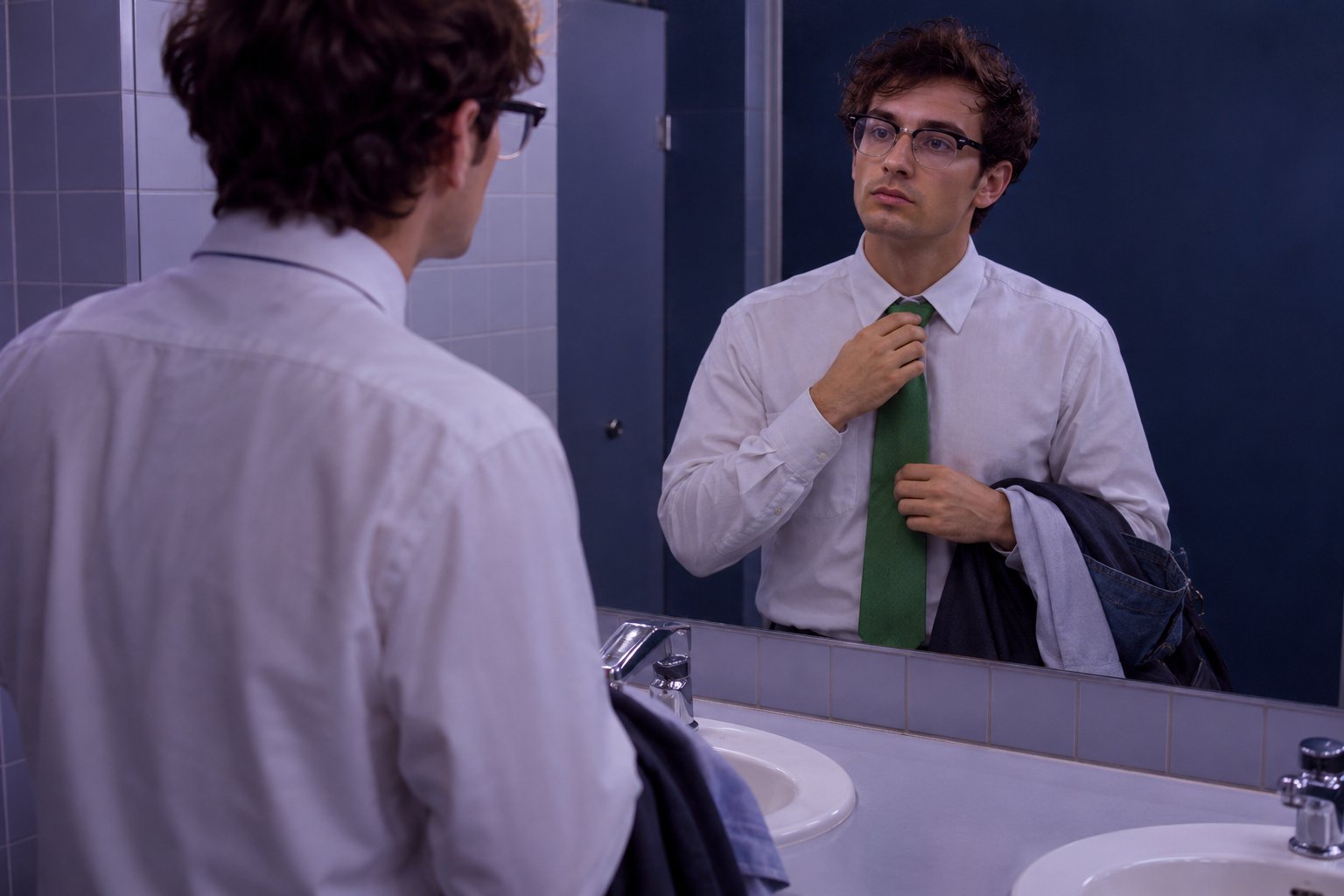 A slim man in a bright green tie and glasses adjusts his tie at a washroom mirror in a 1990s office building.
