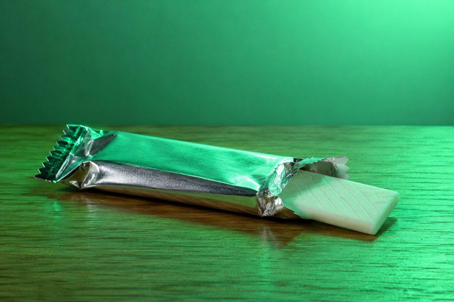 A pack of white chewing gum with one stick sliding out on a wooden table in green light