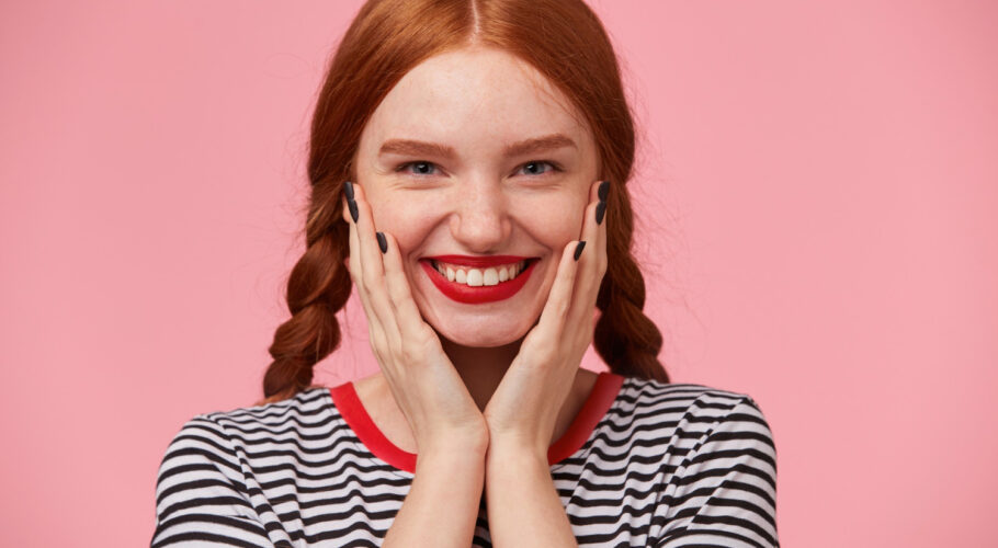 close up glad joyful beautiful red haired girl with two braids keeps hands near her face smiling rosily