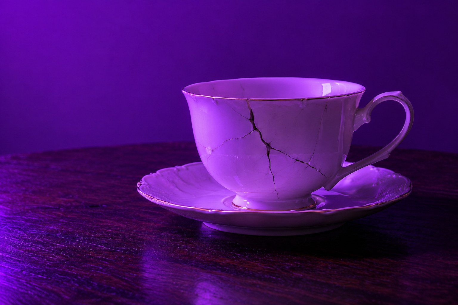 a cracked white porcelain teacup on a saucer on a dark wooden table photographed in a studio