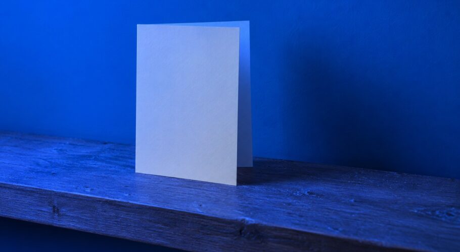 A lone blank greeting card standing on a dusty wooden shelf in blue light