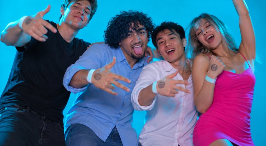 Four young clubbers with wristband stamps dancing and grinning against a plain cyan studio backdrop