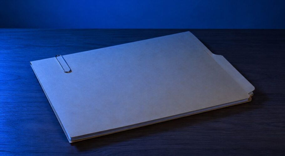 A closed manila folder with a paper clip on a wooden desk under blue light