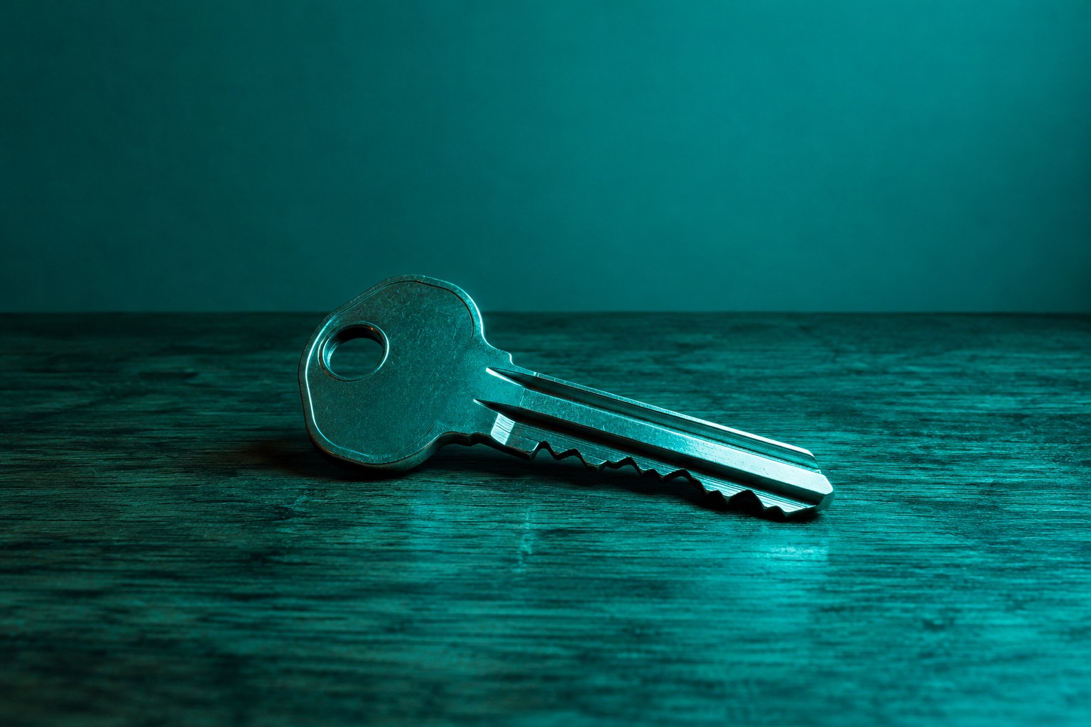 A plain metal house key lying on a wooden table in teal light
