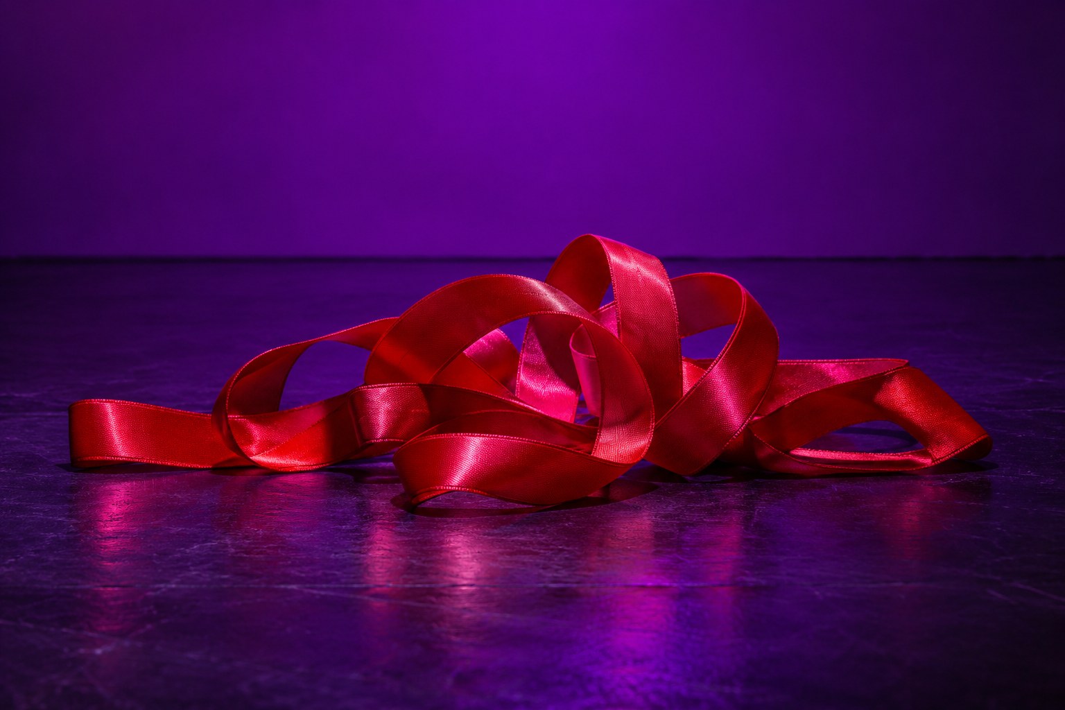 A tangled coil of red satin ribbon on a dark stage floor under purple light