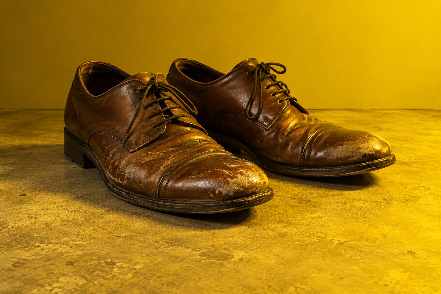 A pair of scuffed brown leather dress shoes on concrete in yellow light