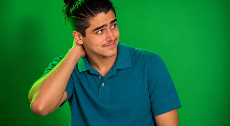 Shy young man with a topknot in a teal polo rubbing the back of his neck and glancing sideways against a plain green studio backdrop