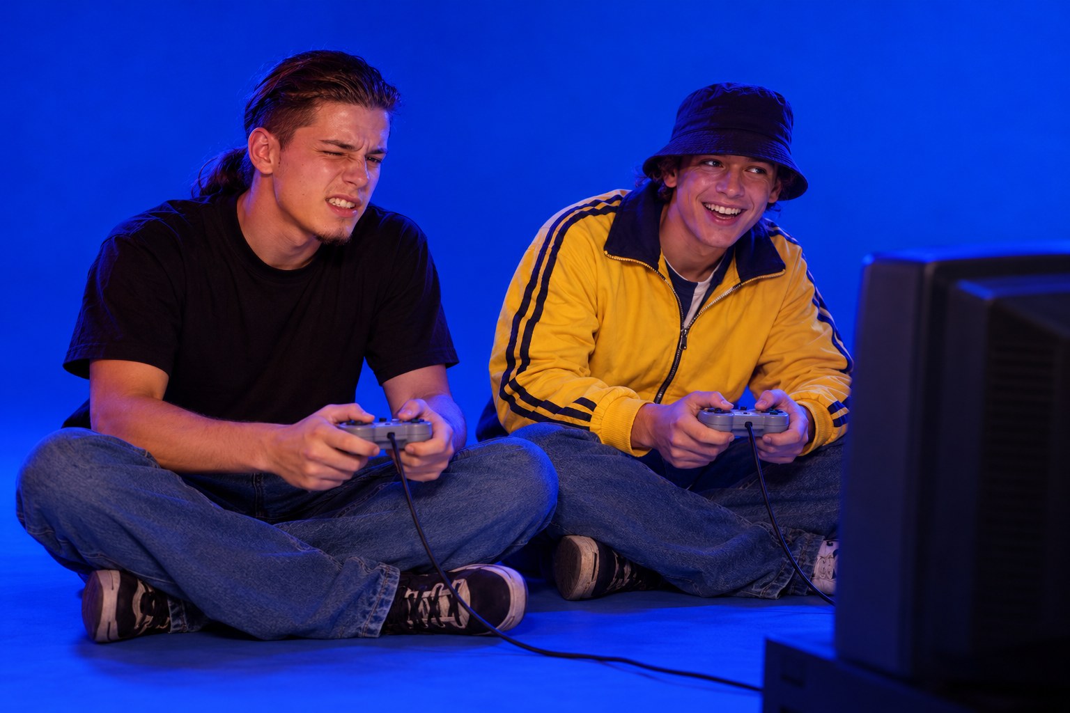 A long-haired young man and a young man in a bucket hat sit cross-legged on a bedroom floor in front of a CRT television, gripping grey Playstation controllers, a long-haired young man wincing as a young man in a bucket hat grins in 1990s Australia