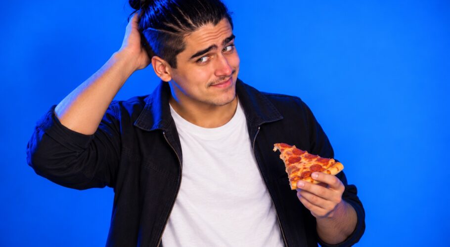Young man with a topknot in a white tee and dark jacket scratching his head and holding a slice of pizza against a plain blue studio backdrop